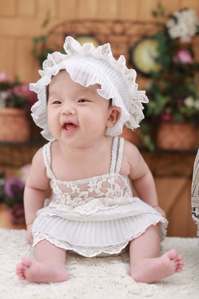 Cute baby girl wearing a lace dress and hat, smiling happily indoors.