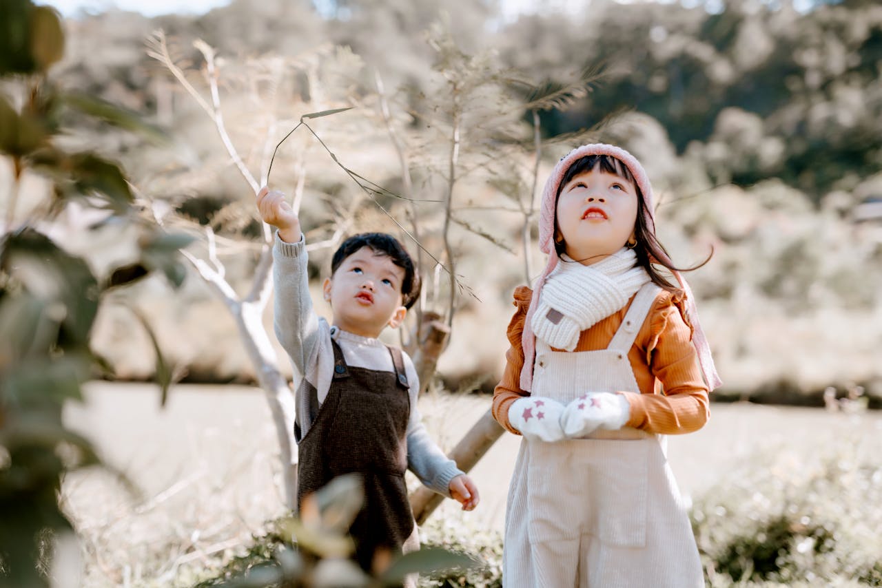services-04 Attentive Asian boy and girl looking up while standing in countryside on grassy terrain with green foliage against blurred background